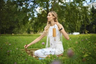 A woman sits calmly on a soft grassy area, softly striking a singing bowl while surrounded by