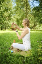 A woman sits cross-legged on a mat in a lush green field, playing a singing bowl with a mallet.