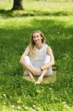 A barefoot woman sits on a mat in a vibrant green park, enjoying the sunny weather. She has long