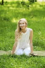 A woman with long hair sits on a yoga mat in a vibrant green park. She wears a light outfit and