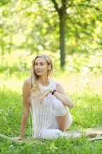 A woman with long hair sits elegantly in a vibrant green park. Dressed in white, she poses