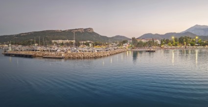 Kemer marina at dusk, filled with multiple docked boats. Lights from the harbor and hotels on the