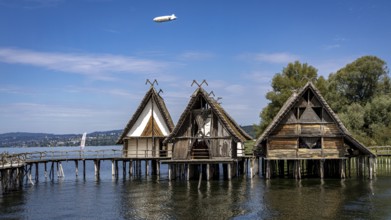 Three wooden pile dwellings with pointed roofs hover over calm waters, pile dwellings