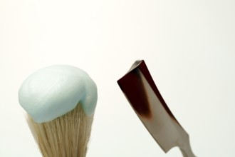 Shaving brush with light blue foam and straight razor on white background, getting ready for