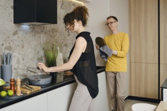A woman stirs a pot on the stove while a a man wipes the washed dishes and watches her in a stylish