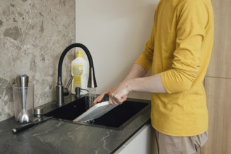 A person is cleaning a chopping board under running water in a sleek modern kitchen. The individual