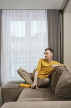 A dreamy man dressed in a yellow shirt and beige pants sits comfortably on a sofa, casually resting