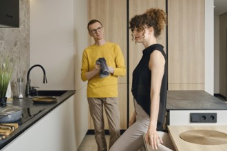 In a stylish kitchen, a man in a yellow shirt looks thoughtfully at a woman in a black top as she