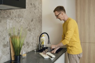 A man in a yellow long-sleeve shirt is washing utensils at a sleek kitchen sink. Natural light