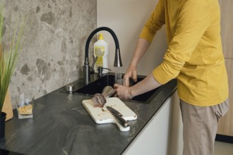 A person wearing a yellow shirt is washing utensils and a cutting board in a dark kitchen sink. The