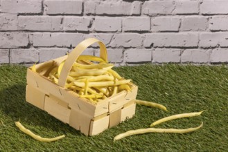 Wooden basket with yellow beans on grass in front of a brick wall