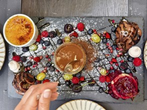Hand with spoon tasting a dessert, surrounded by artfully arranged berries and chocolate Lower