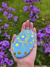 Hand holding blue stone with gold lettering and dots, purple flowers in the background Lower