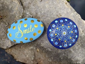Two painted stones on rocks, one with 'Smile', one with a mandala pattern in blue and gold Lower