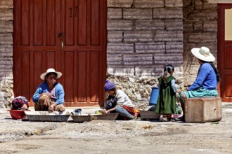 Women and children sit in front of a stone house, busy with housework. The sun shines brightly,