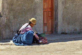 A woman in traditional dress sits on the street in front of a wall in the sun, Traditionally