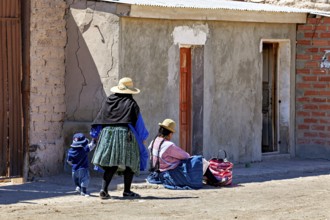 Woman and child in traditional dress standing in front of a brick house on a sunlit street,