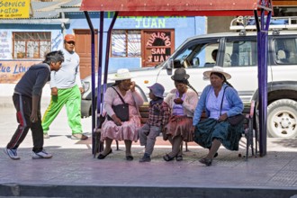 A group of people in traditional dress sit on a bench surrounded by shops and a car, people at a