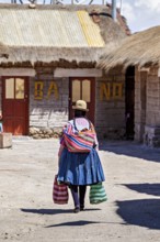 A woman in colourful traditional dress carries bags in a village with rustic buildings, Traditional