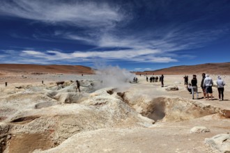 People standing in a desert landscape with a smoking geyser and blue sky, The mud volcanoes and