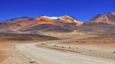 A winding sandy road through a dry landscape with reddish mountain peaks and blue skies, The