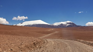 Dusty road leads through a dry desert with snow-capped mountains in clear daylight, The landscape