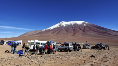 Groups of people and vehicles in a desert landscape with a snow-covered mountain under a blue sky,