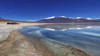 Panoramic view of a lake with snow-capped peaks, people and vehicles on the shore, The Laguna
