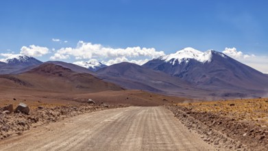 A straight, unpaved road leads through a barren mountain desert landscape on a sunny day, The