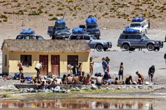 Group of people and cars in front of a small building in a desert landscape, The Aguas Termales de