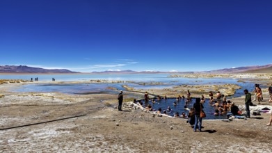 People bathing in a small lake in a desert landscape under a bright blue sky, The Aguas Termales de