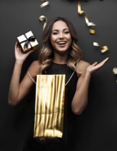 Smiling woman holding a small gift box and golden shopping bag with festive gold ribbons on a black