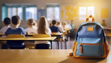 A brightly colored school backpack is placed on a desk in the foreground, with students seated in a