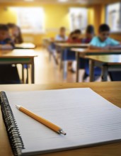 A notebook with a pencil rests on a classroom desk, with students working in the background,