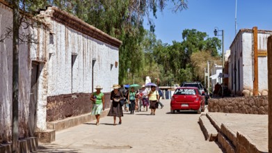 Pedestrians in summer clothes walk along a street with white buildings and a red car, The town of