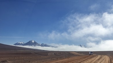 A dusty road leads through a remote desert landscape, bordered by mountains and clouds under a