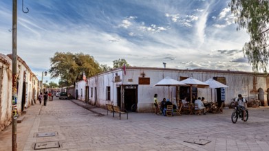 People move along a street with old buildings under partly cloudy skies and cyclists, The town of