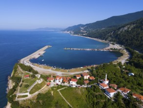 Aerial view of a coastal town with red roofs surrounded by green hills and calm blue sea, aerial