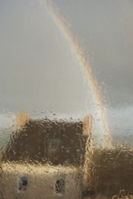 A house seen through a rain-soaked window with a rainbow arching across a gray sky, Old house