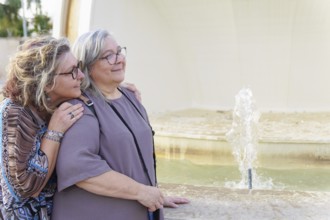 Two happy senior women hugging each other near a fountain in a city park, enjoying a sunny day