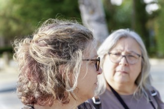 Two senior women wearing eyeglasses are enjoying a conversation on a sunny day in a park