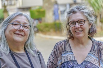 Two mature women with gray hair and glasses are smiling outdoors, possibly enjoying a leisurely day