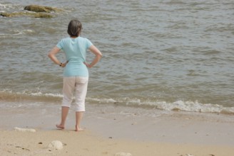 A woman stands on a sandy beach looking at the ocean waves, with rocks in the background in Royan