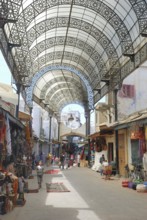 A vibrant market street with ornate metal canopy, bustling with people and colorful merchandise.