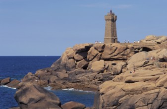 A lighthouse stands on a rocky coastline with people nearby and a clear blue sky. The granit rose