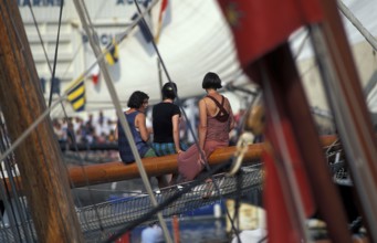 Three women sitting on a sailboat's wooden deck surrounded by nautical flags and ropes. Maritime
