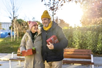 Elderly couple holding christmas presents smiling and walking in a city park during a sunny winter