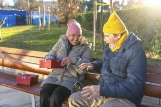 Elderly man surprising his wife with christmas gifts while sitting on a bench in a city park