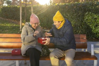 Elderly man surprising his wife with a festive christmas gift while sitting together on a park
