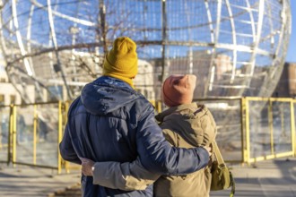 Elderly couple embracing while admiring large christmas ball decorations in a winter cityscape